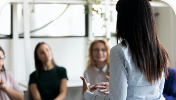 Image or woman standing in foreground and speaking to a group of seated women in the back
