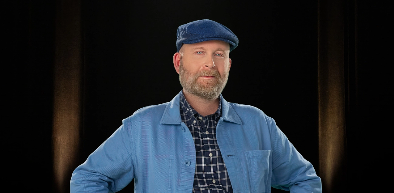 An older man with a beard wearing a driving cap and blue shirt smiling with his arms crossed.