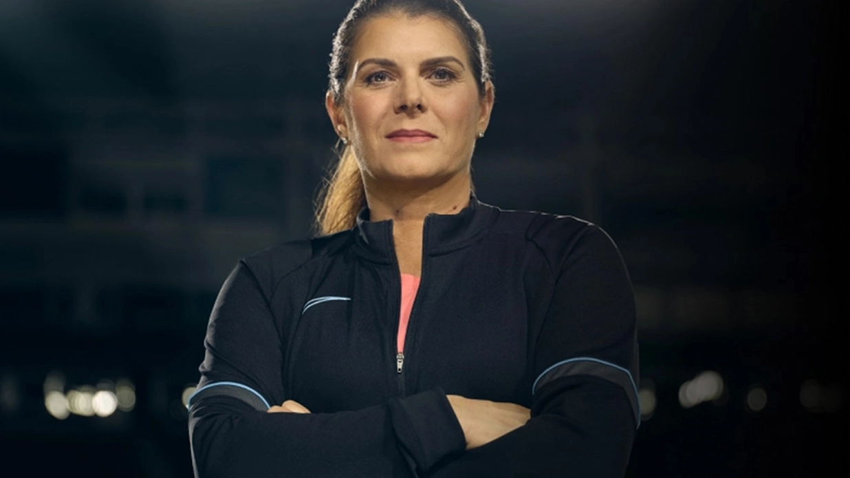 Mia Hamm wearing a black jacket over a pink shirt, looking confidently into the camera with her arms folded.