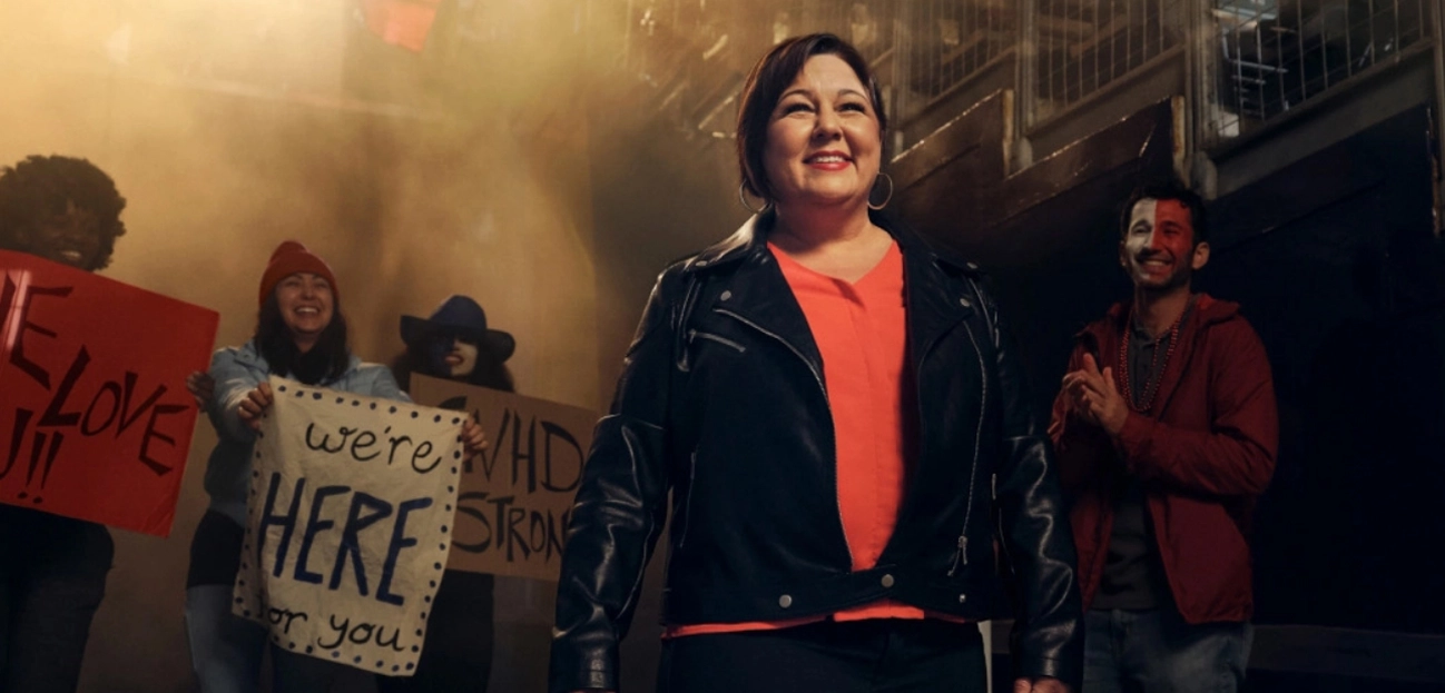 A middle-aged woman with short, brown hair wearing a coral shirt and black leather jacket smiles at the camera.