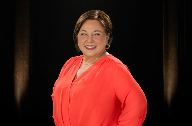 A middle-aged woman with brown hair and a coral shirt smiles at the camera with a hand on her hip.