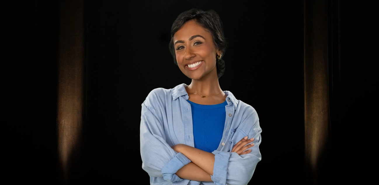 A young woman with short brown hair, wearing a blue shirt and white pants, smiles with her arms crossed.