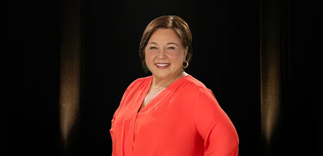 A middle-aged woman with brown hair and a coral shirt smiles at the camera with a hand on her hip.
